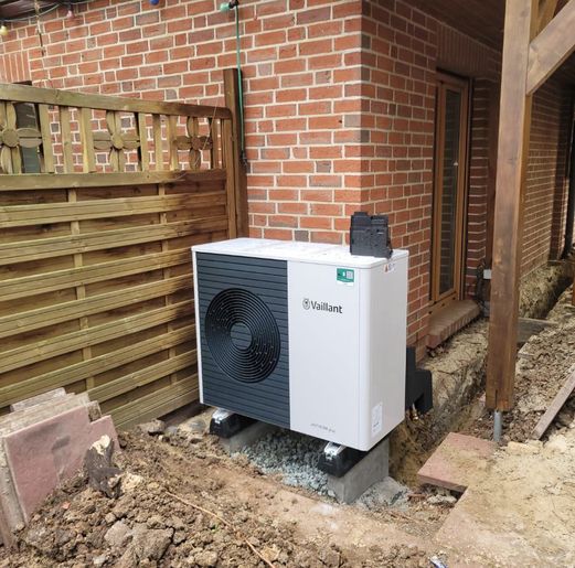 Eine weiße Wärmepumpe der Marke Vaillant, aufgestellt auf einem Betonfundament neben einer Ziegelwand und einem Holzzaun im Gartenbereich